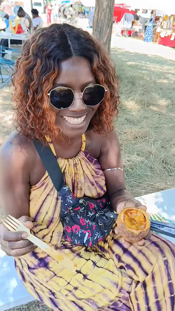 Femme souriante en robe tie-dye jaune et violette avec lunettes de soleil, tenant un fruit de maad ouvert, assise en plein air lors d'un marché ensoleillé.